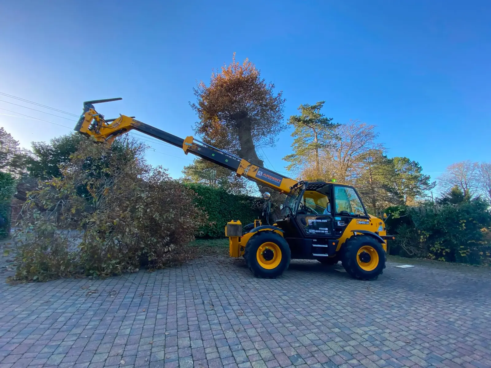 Telehandler clearing storm-damaged hedge and tree debris from a driveway